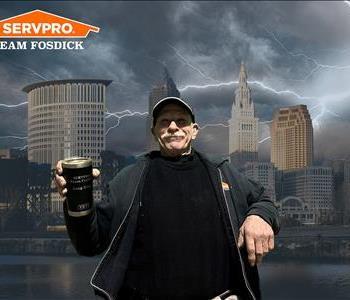 Man wearing a har with a mustache smiling holding a coffee mug in front of a stormy Cleveland background